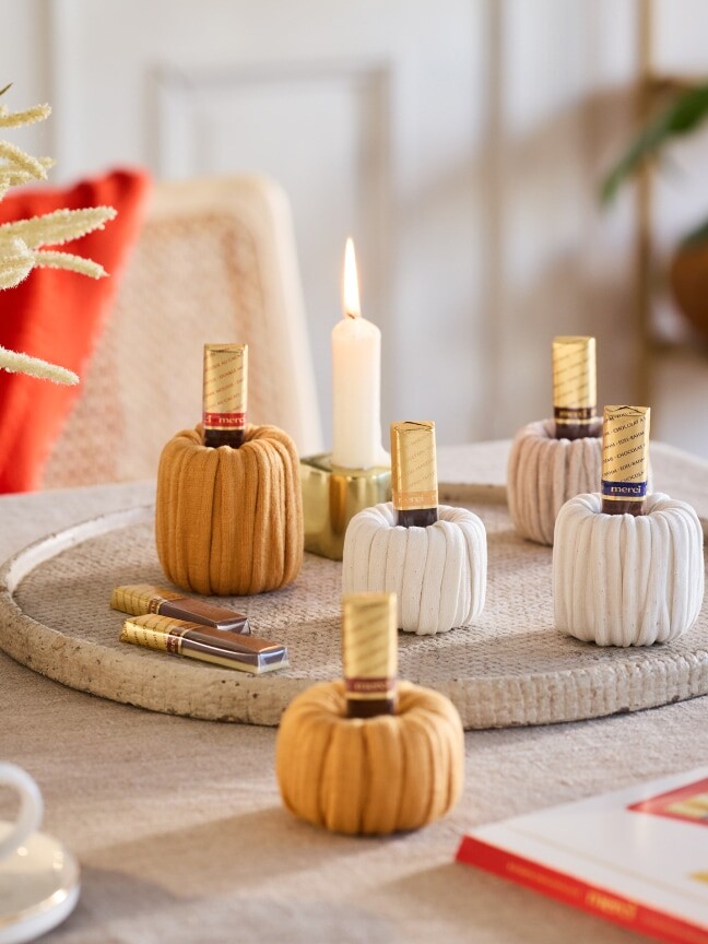 Die DIY-Dekoidee zeigt selbst gebastelte Kürbisse aus Garn mit merci Schokolade als Stiel, die auf einem herbstlich dekorierten Esstisch stehen.